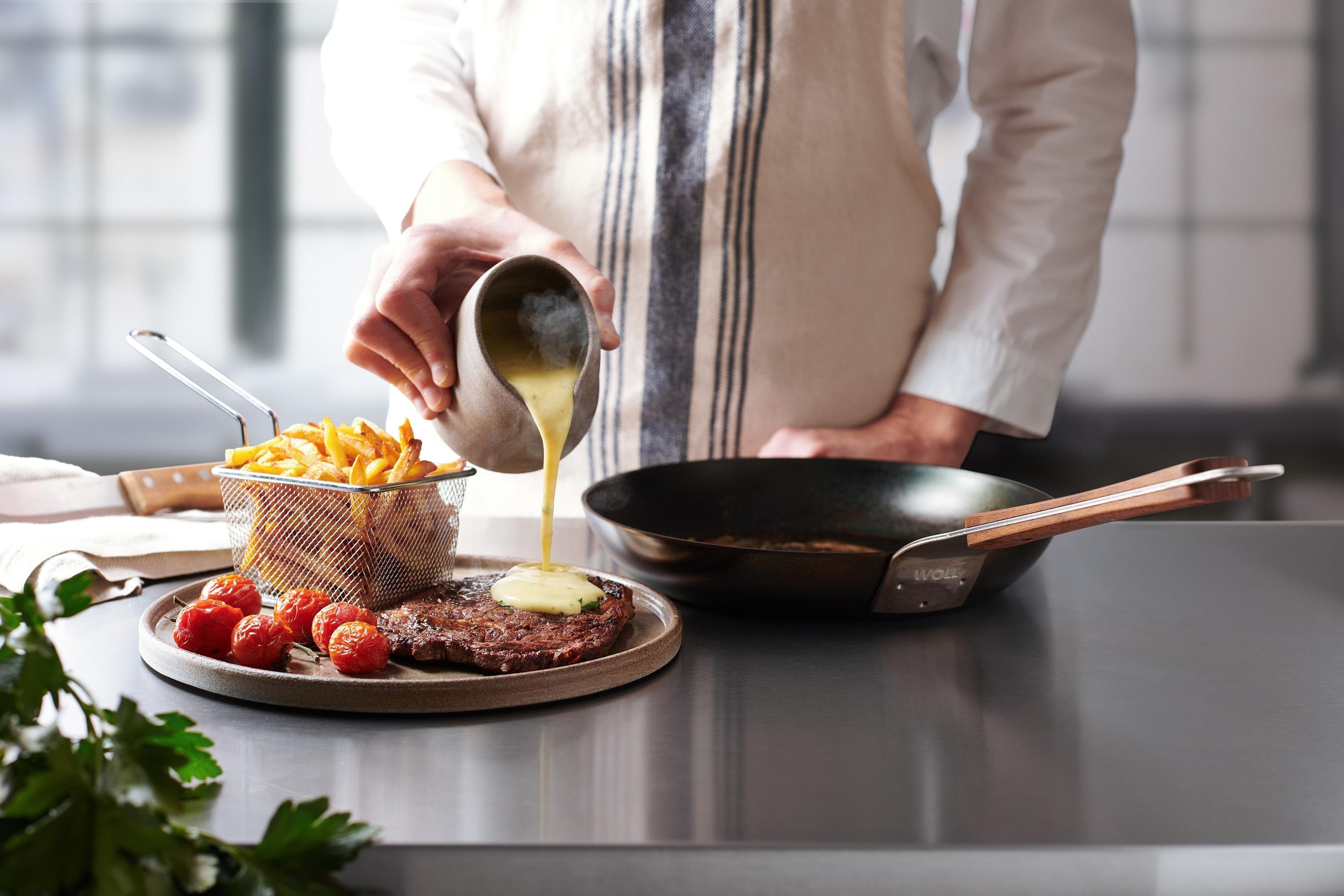 Auf einem Teller liegt ein frisches, angebratenes Steak, das mit Soße übergossen wird. Daneben steht ein kleiner Korb mit Pommes und liegen angebratene Cherrytomaten. Auf der Arbeitsplatte steht eine runde Carbon Steel Pfanne mit festem Holzstiel.
