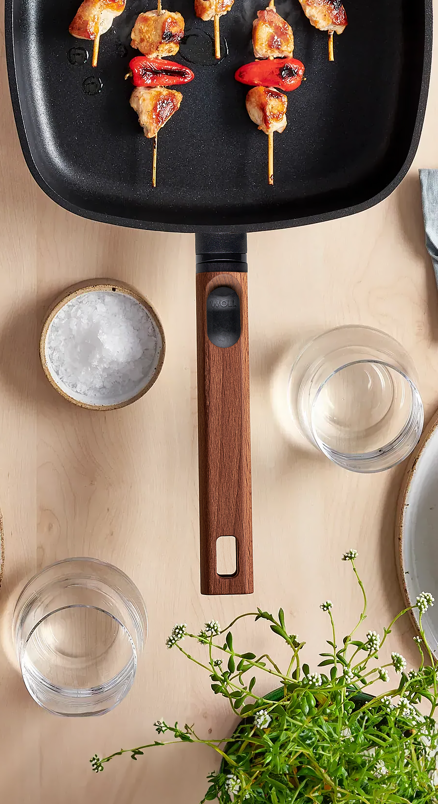 Auf dem Tisch steht eine viereckige Pfanne mit abnehmbarem Stiel aus der Serie Eco Logic QXR, die frisch angebratene Fleischspieße enthält. Drumherum stehen Teller, Gläser, Gewürze und eine Pflanze.