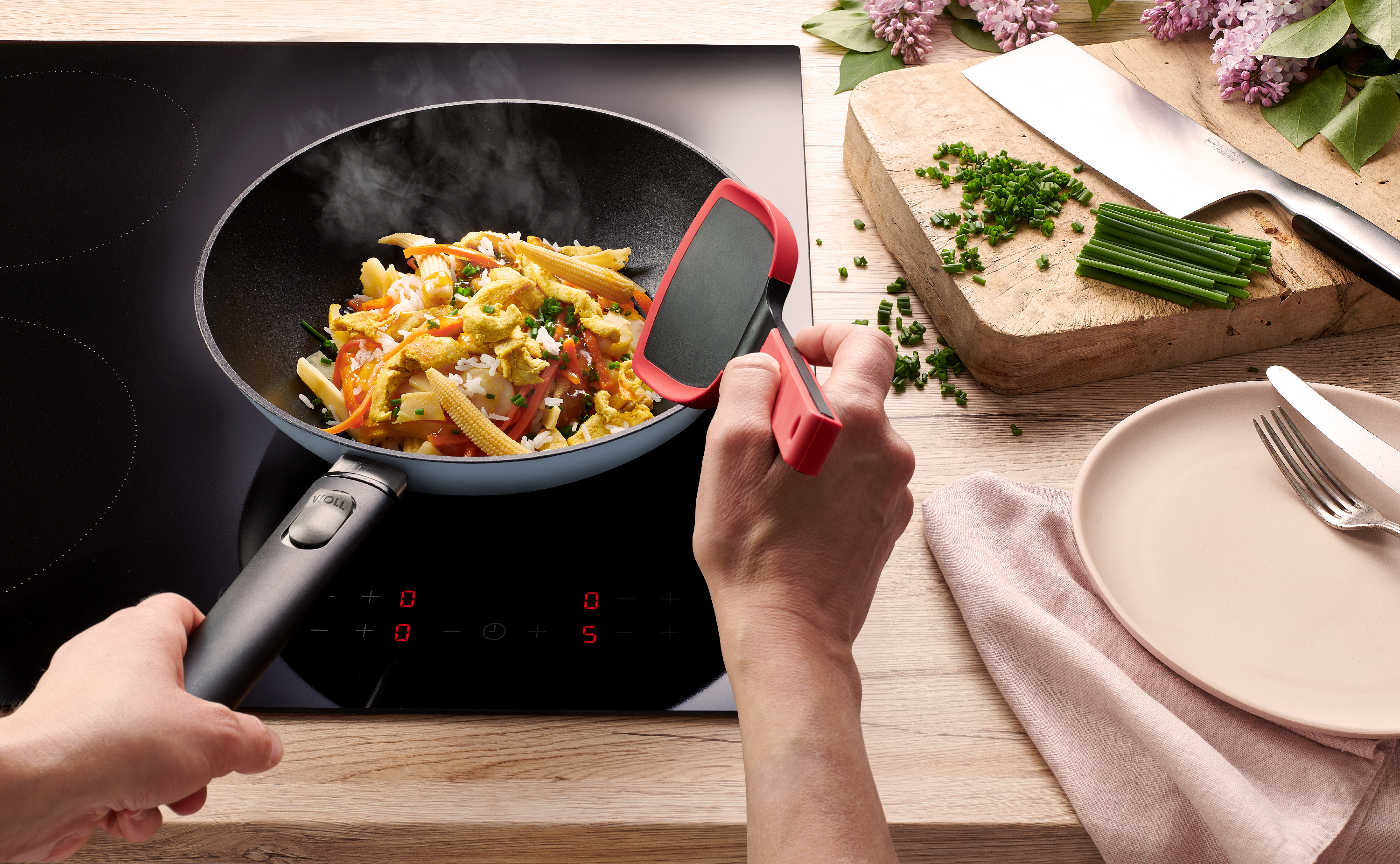 Eine runde, himmelblaue Ecolour Wokpfanne steht auf dem Herd, mit einem Cock it wird ein dampfendes Gericht gewendet. Das Edge Chinesisches Kochmesser liegt daneben.