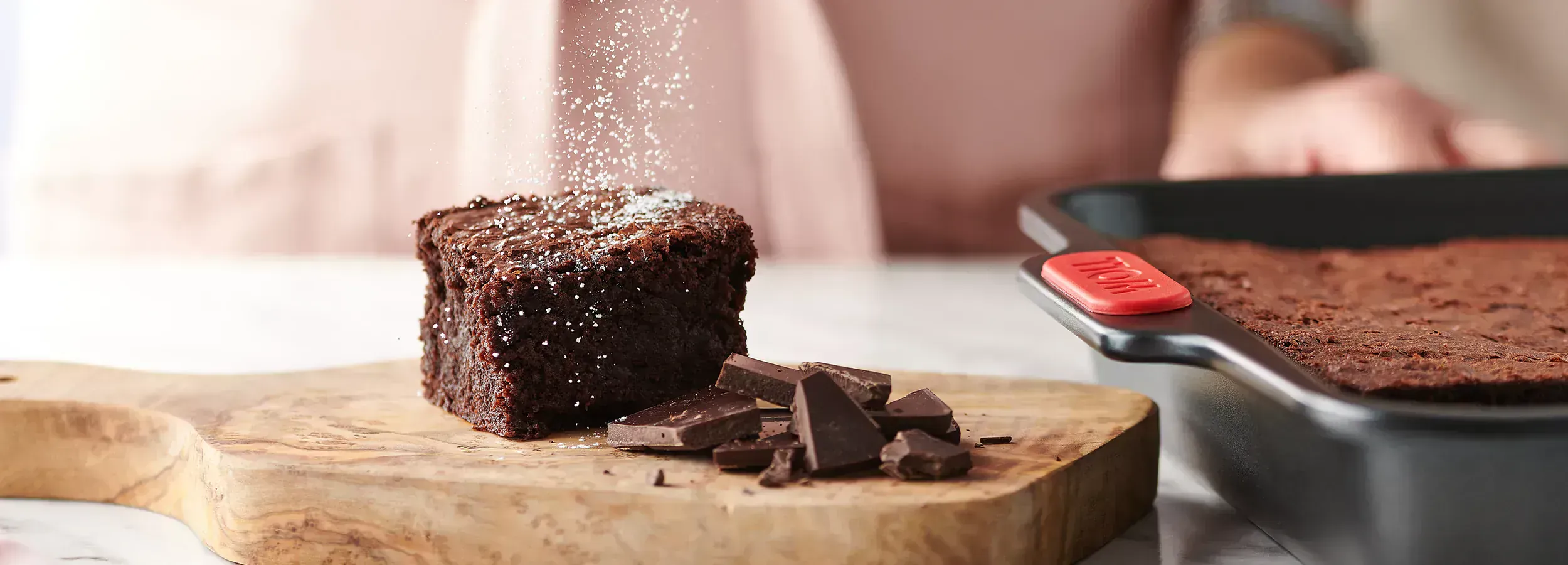 Auf einem Holzbrett liegt ein frisch gebackenes Stück Schokoladenkuchen, das mit Puderzucker bestäubt ist. Schokoladenstückchen auf dem Holzbrett dienen als Dekoration. Daneben steht die rechteckige Backform der Let's Bake!-Serie, die mit weiterem Schokol