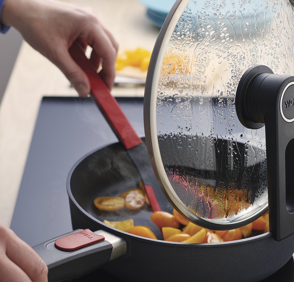 In der runden Diamond Lite Pfanne wird ein Gericht mit einem roten Cock it Pfannenwender umgerührt. Der Smart Lid steht auf der Pfannenkante.