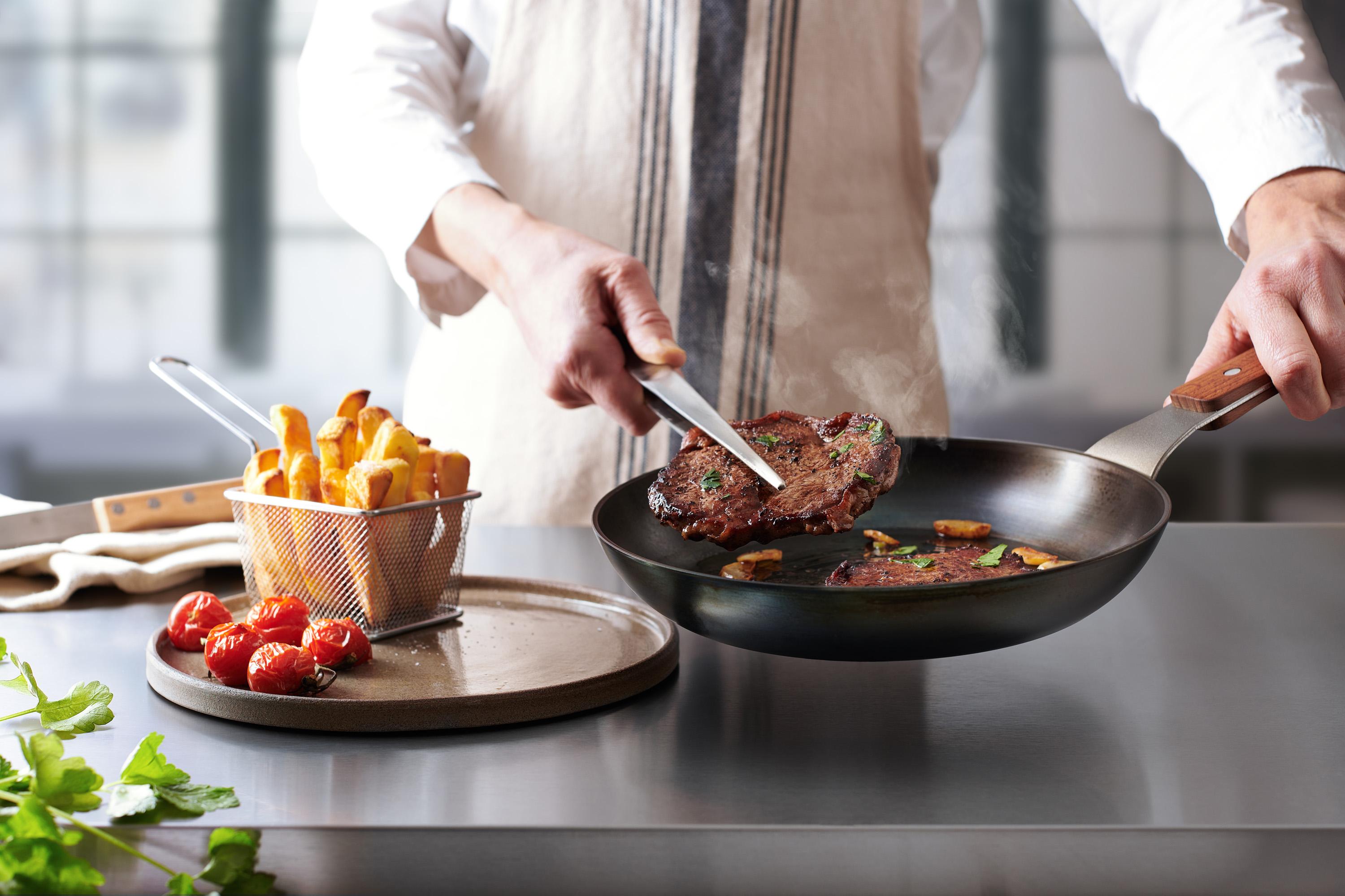 In einer runden Carbon Steel Pfanne befinden sich angebratene Steaks. Ein Steak wird auf einem Teller platziert.