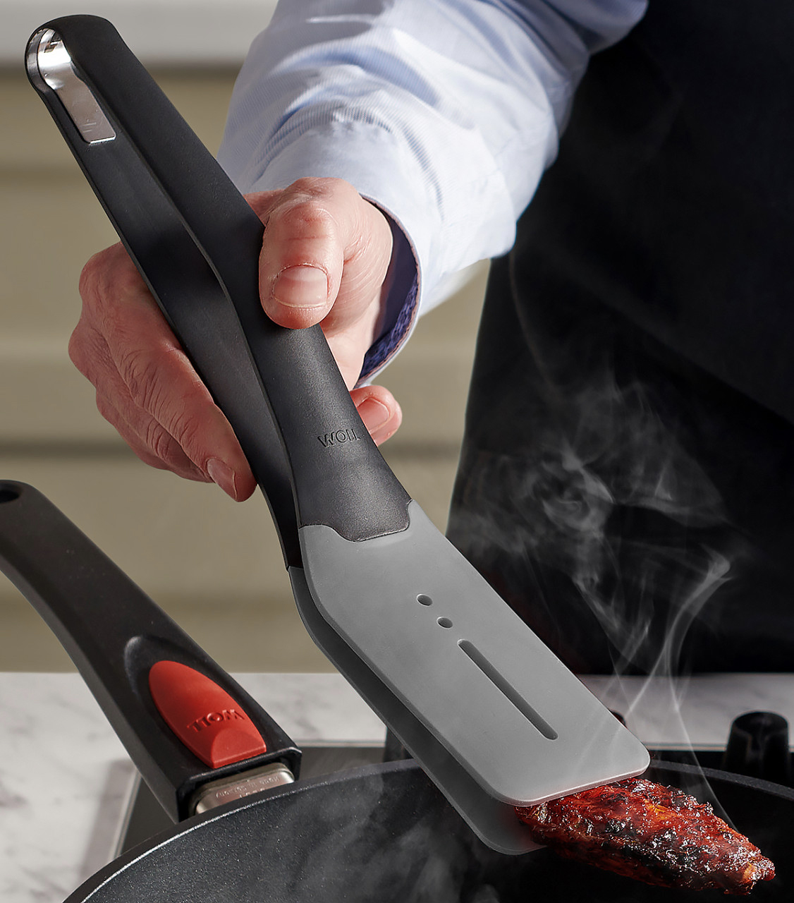 Ein dampfendes, angebratenes Stück Fleisch wird mithilfe der grauen Cock it Küchenzange in der heißen Pfanne gewendet.