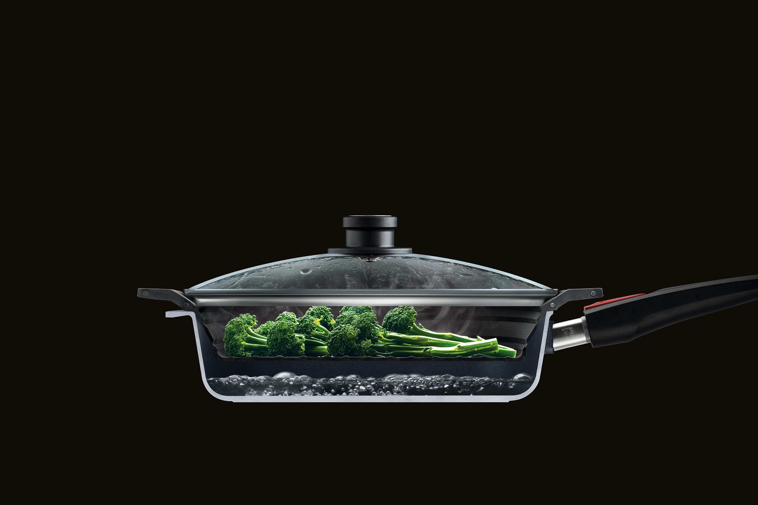 Im Querschnitt ist eine halbierte Wok Pfanne mit Glasdeckel zu sehen, in der Brokkoli mit dem Steam it multifunktionalen Silikoneinsatz gedämpft wird. 