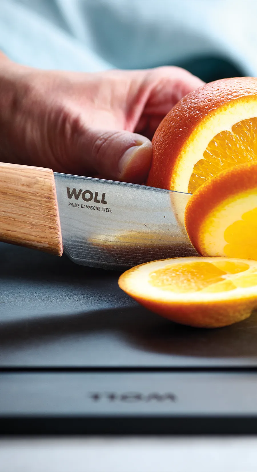 Eine Orange wird auf einem Schneidbrett mit einem Prime Edge Messer in Scheiben geschnitten, während daneben zwei ganze Orangen liegen.