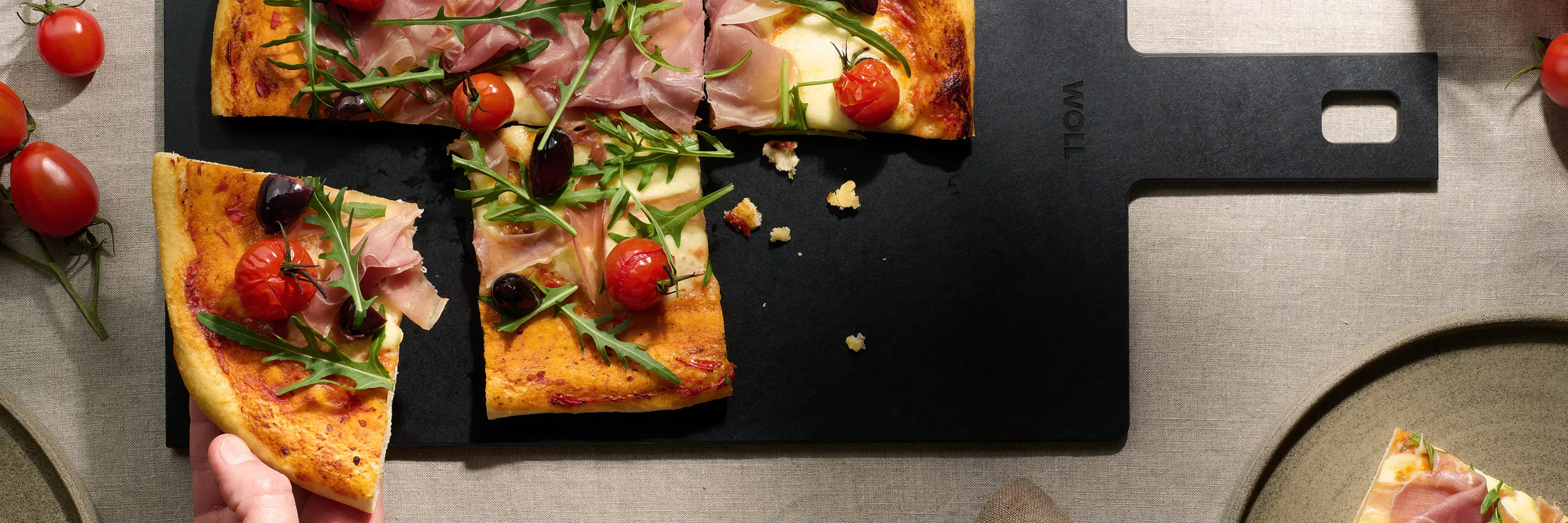 Auf dem Schneidebrett mit Griff der Serie „Cut & Serve” liegen frisch gebackene Pizzastücke. Außen herum stehen ein Glas, ein Teller und Tomaten als Dekoration.