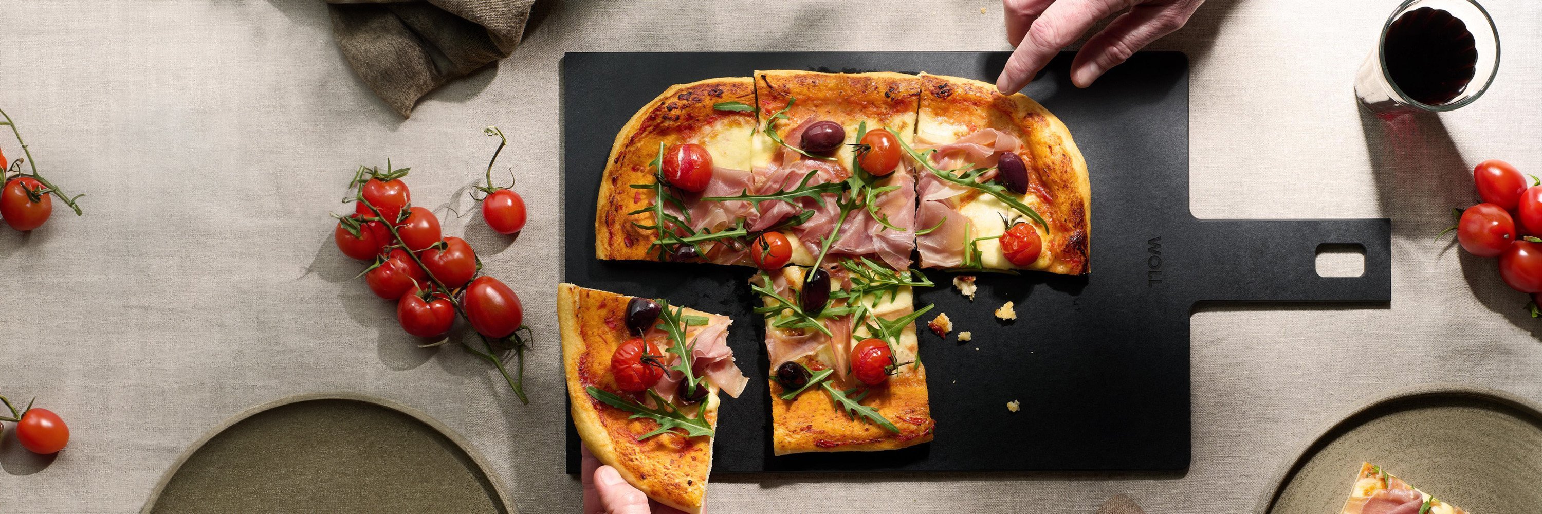 Auf dem Schneidebrett mit Griff der Serie „Cut & Serve” liegen frisch gebackene Pizzastücke. Außen herum stehen ein Glas, Teller und Tomaten als Dekoration.