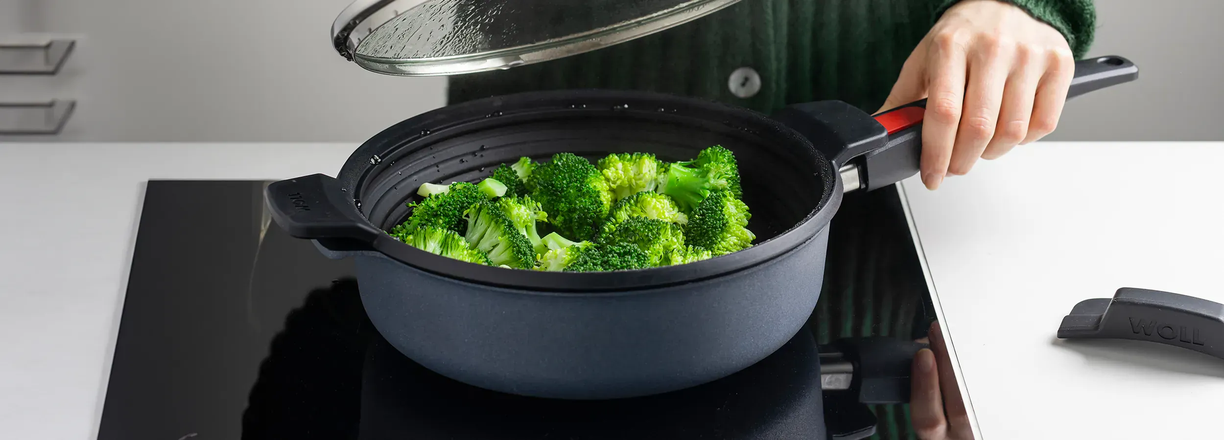 In der Nahaufnahme ist der runde, multifunktionale Silikoneinsatz der Serie Steam it zu sehen, der im Topf platziert ist, in dem der frische Brokkoli gedämpft wird. Der Glasdeckel wurde entfernt. Auf der Arbeitsplatte liegt ein Silikongriffüberzug bereit,