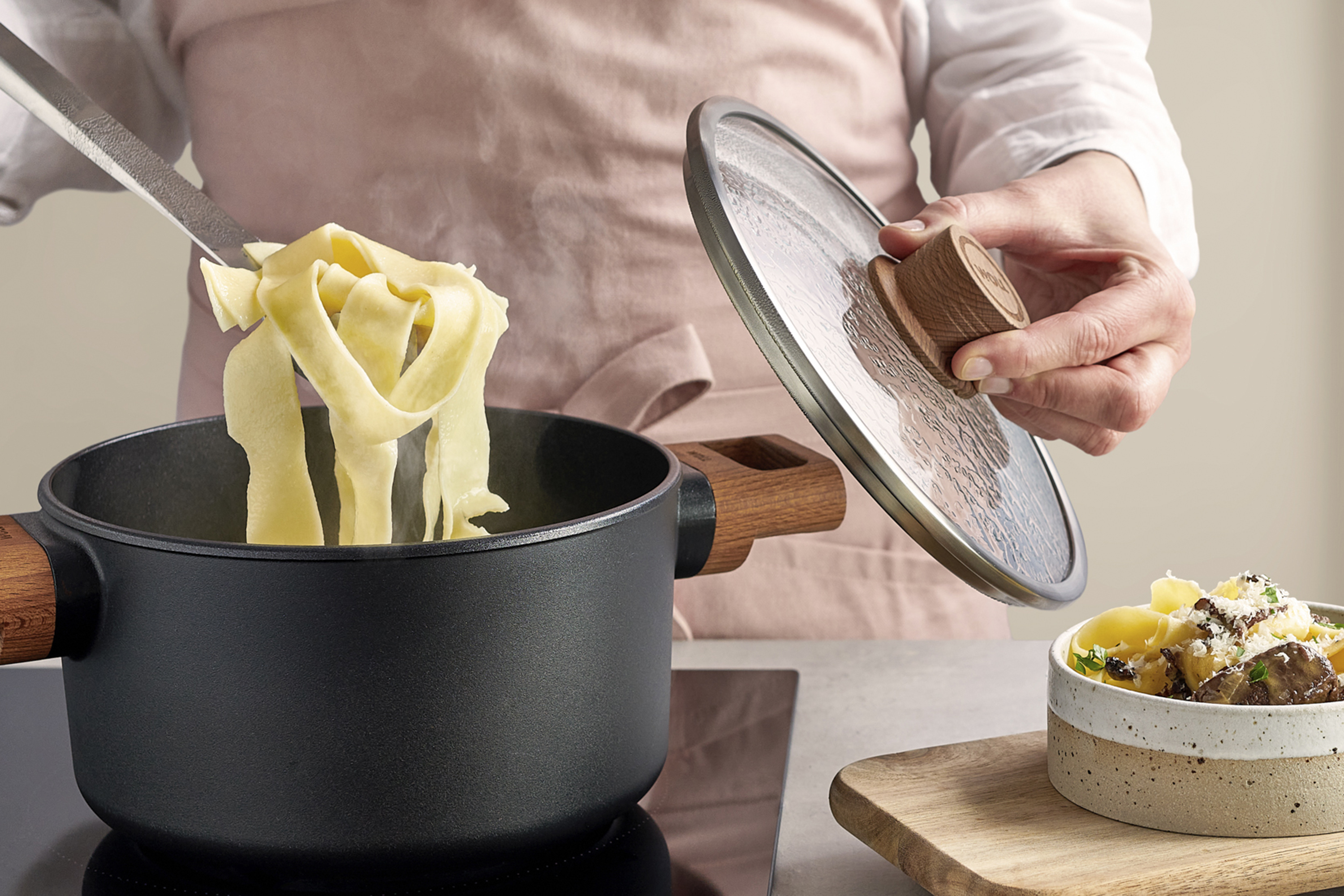 In einem runden Topf der Serie Eco Logic QXR  mit Seitengriffen aus Holz werden die frisch gekochten Nudeln entnommen, während der Deckel mit einer Hand festgehalten wird. Rechts daneben steht auf einem Holzbrett ein fertig gemachtes Nudelgericht.