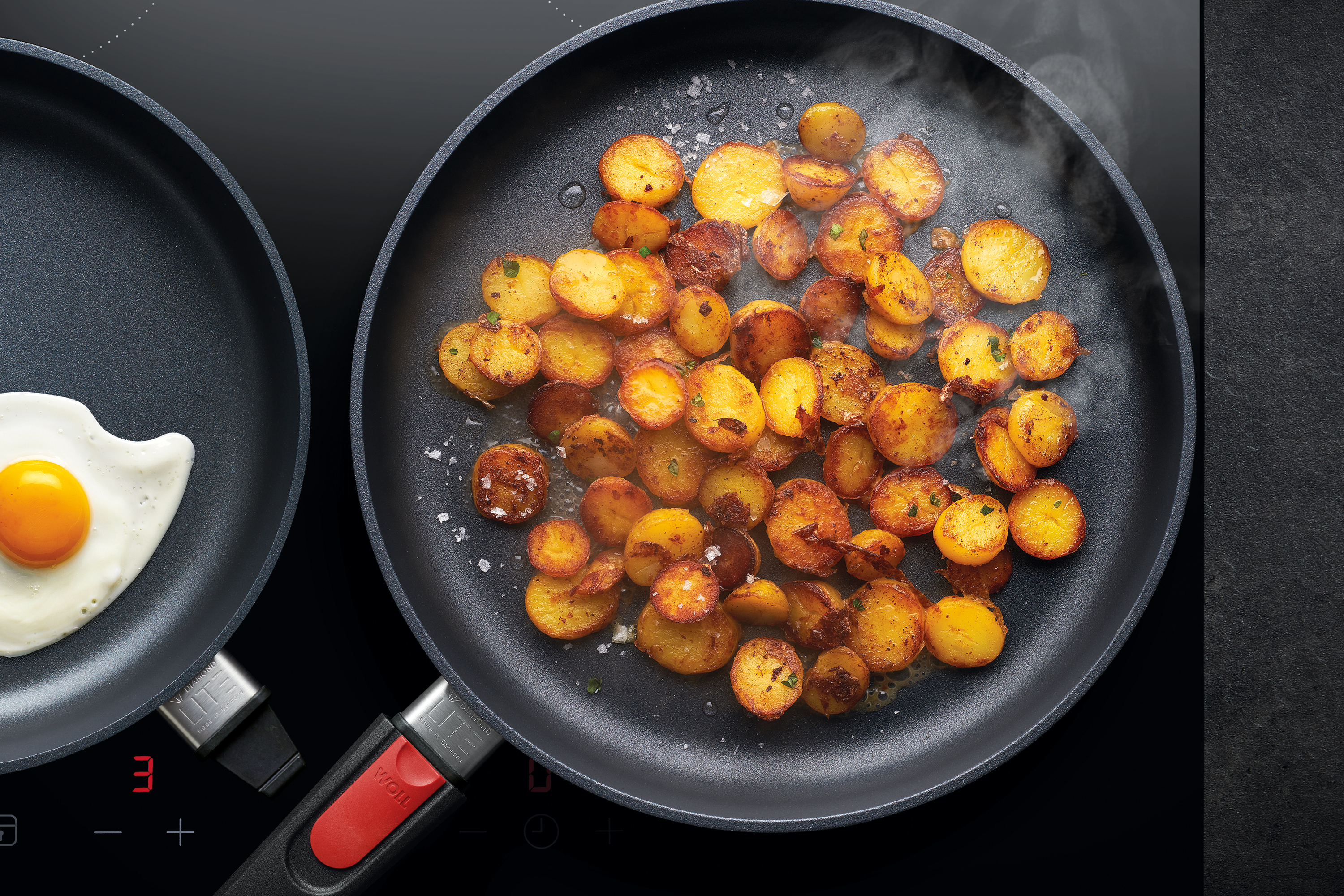 In einer runden Diamond Lite Pfanne werden Bratkartoffeln angebraten, während in einer weiteren Pfanne Spiegeleier zubereitet werden. 