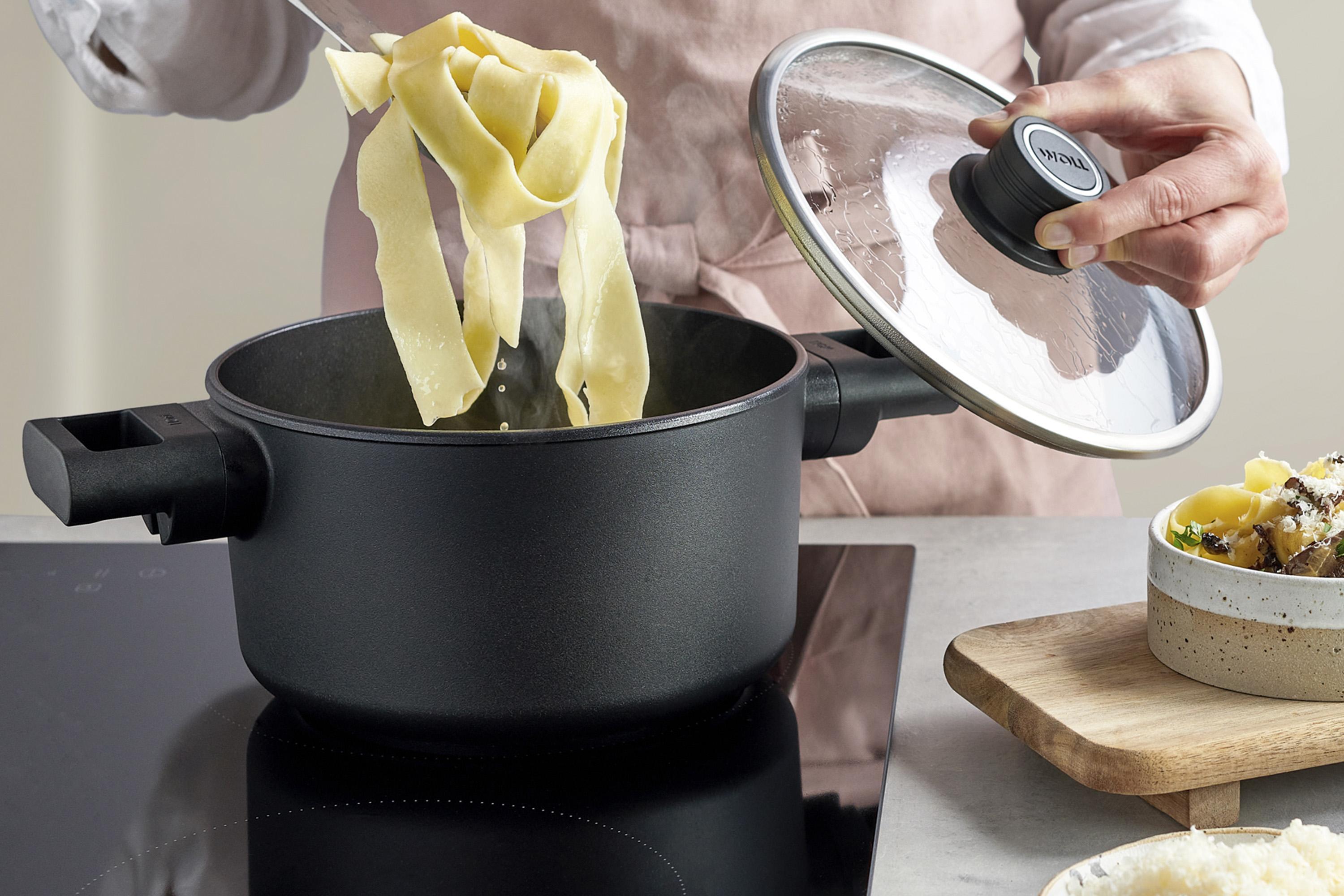 In einem runden Topf der Serie Eco Logic QXR werden die frisch gekochten Nudeln entnommen, während der Deckel mit einer Hand festgehalten wird. Rechts daneben steht auf einem Holzbrett ein fertig gemachtes Nudelgericht.