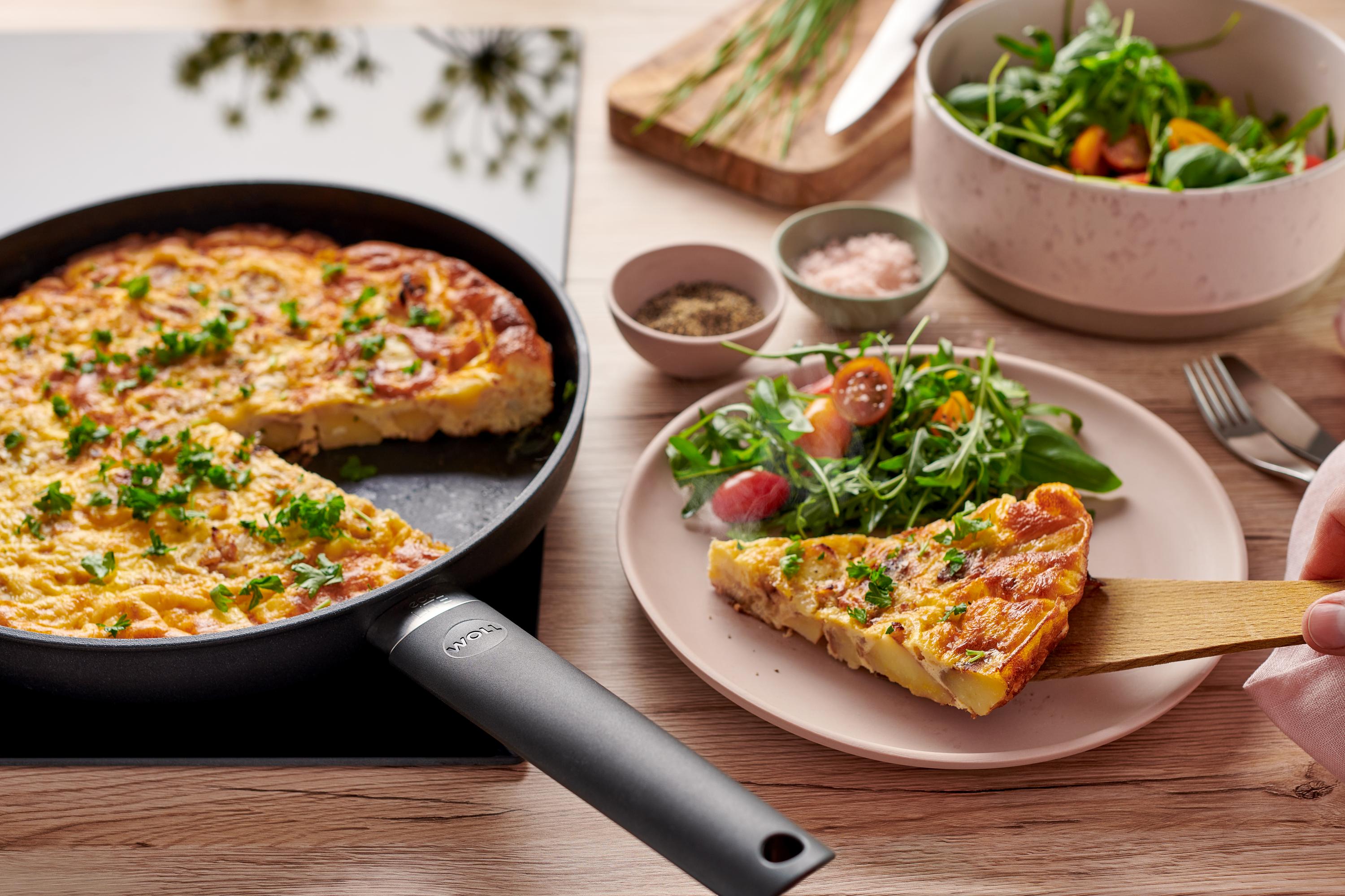 In einer runden Eco Lite QXR Pfanne befindet sich eine frische spanische Tortilla. Ein Stück davon liegt dem Teller.