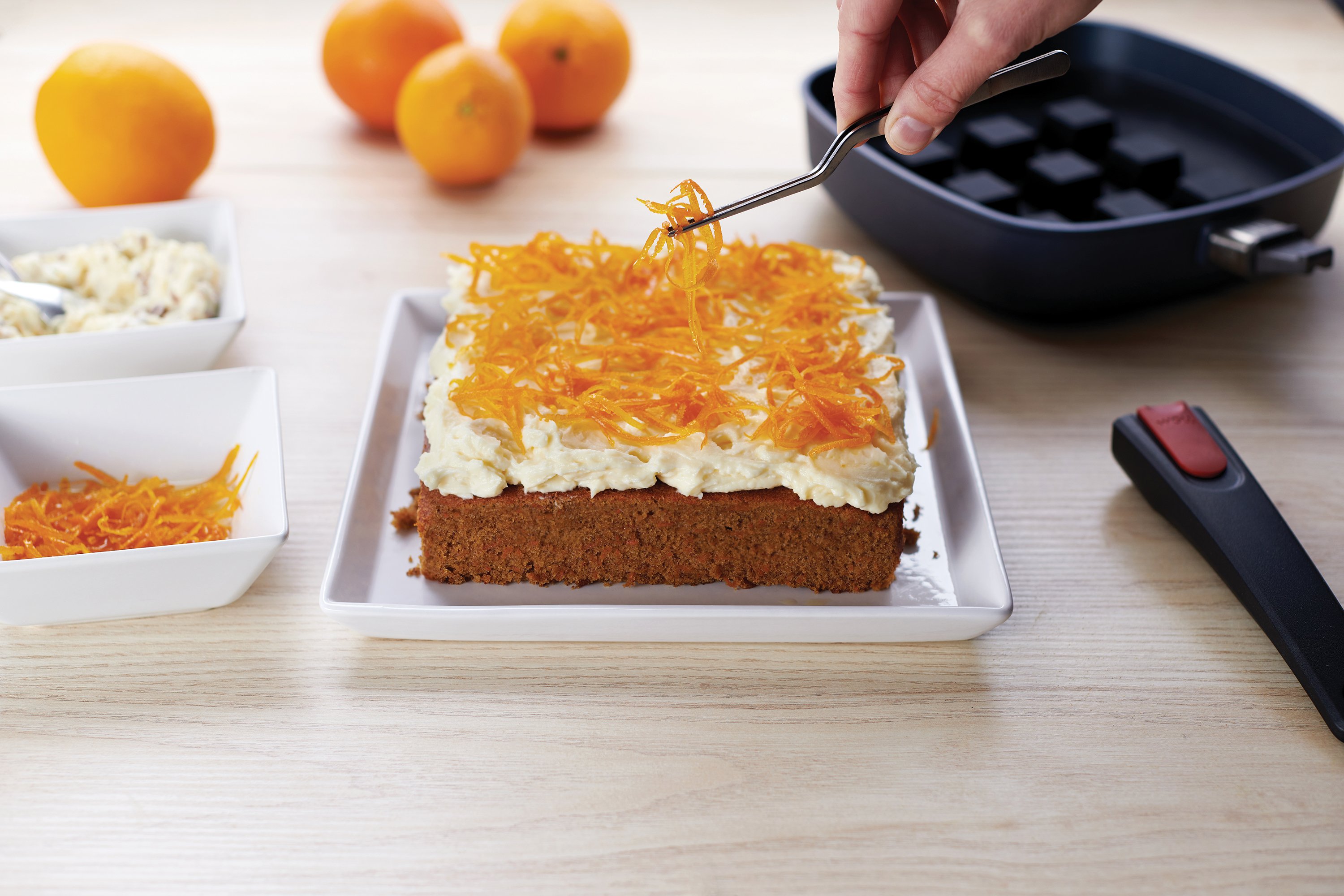 Ein frisch gebackener Kuchen ist mit cremigem Topping und Orangenabrieb garniert. Im Hintergrund ist eine viereckige Pfanne mit Bake it Silikon-Backeinsatz zu sehen.