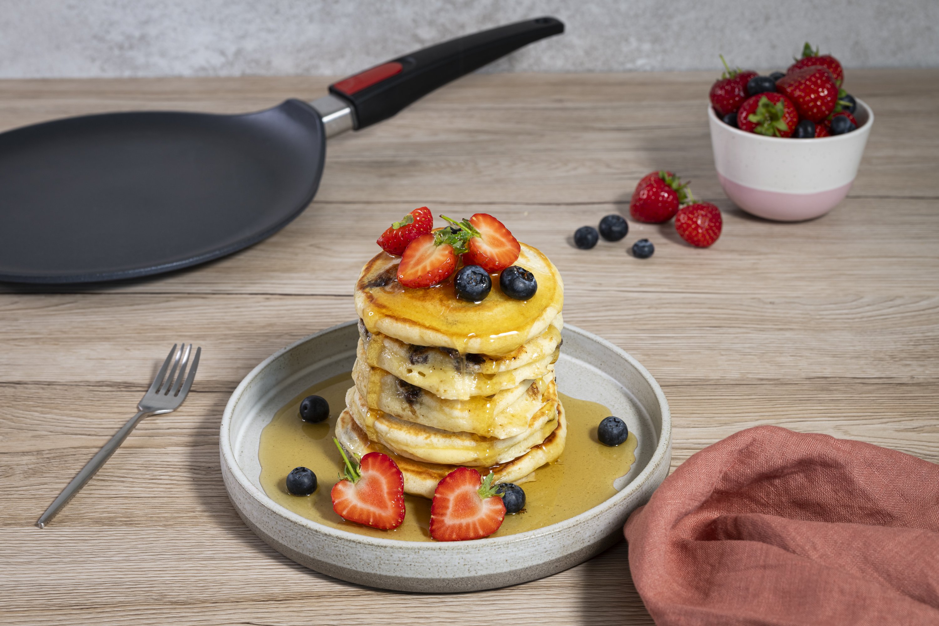 Auf einem Teller wurde ein Stapel frischer American Pancakes angerichtet. Die Pancakes sind mit frischen Beeren garniert und werden mit Ahornsirup übergossen. Im Hintergrund sieht man eine runde Crêpepfanne mit abnehmbarem Stiel.