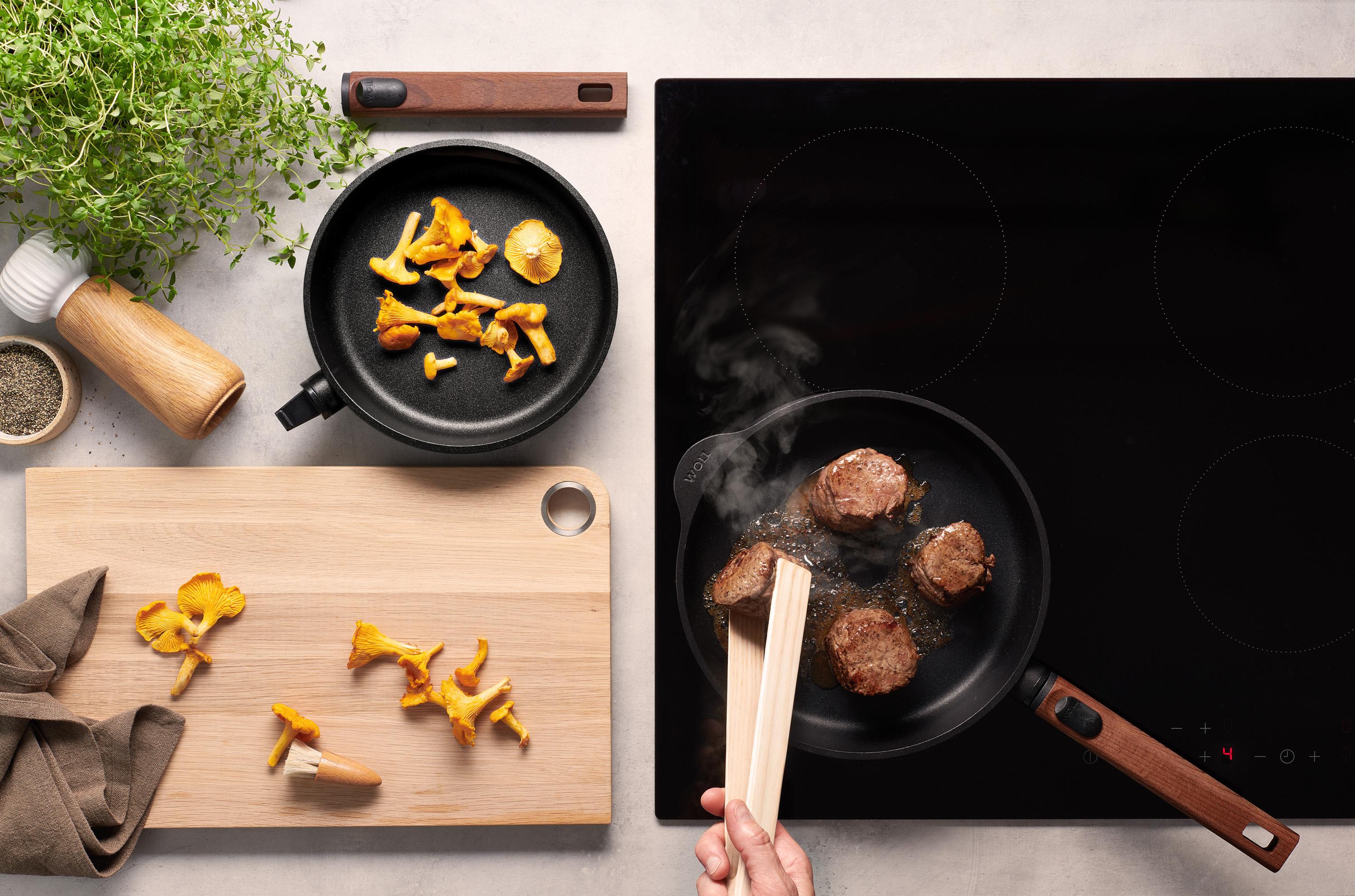 Auf dem Herd steht eine runde, heiße Pfanne der Serie Eco Logic QXR mit abnehmbarem Holzstiel, in der Fleisch angebraten und mit einer Zange gewendet wird. Rechts daneben steht eine weitere Pfanne derselben Serie, deren Holzstiel abgenommen wurde. In der