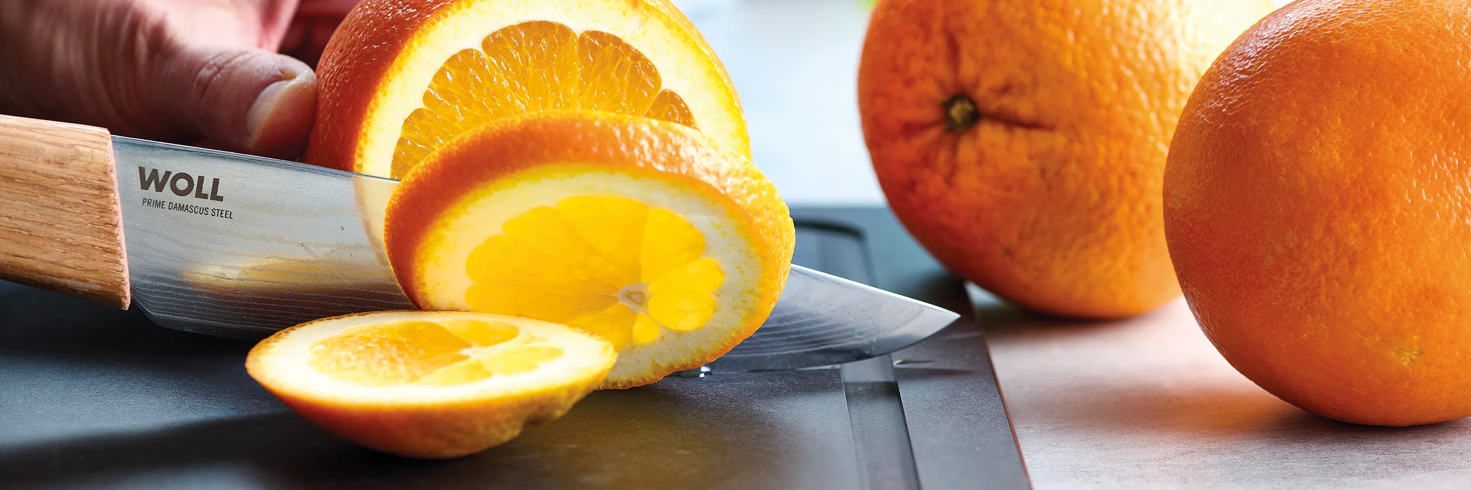 Eine Orange wird auf einem Schneidbrett mit einem Prime Edge Messer in Scheiben geschnitten, während daneben zwei ganze Orangen liegen.