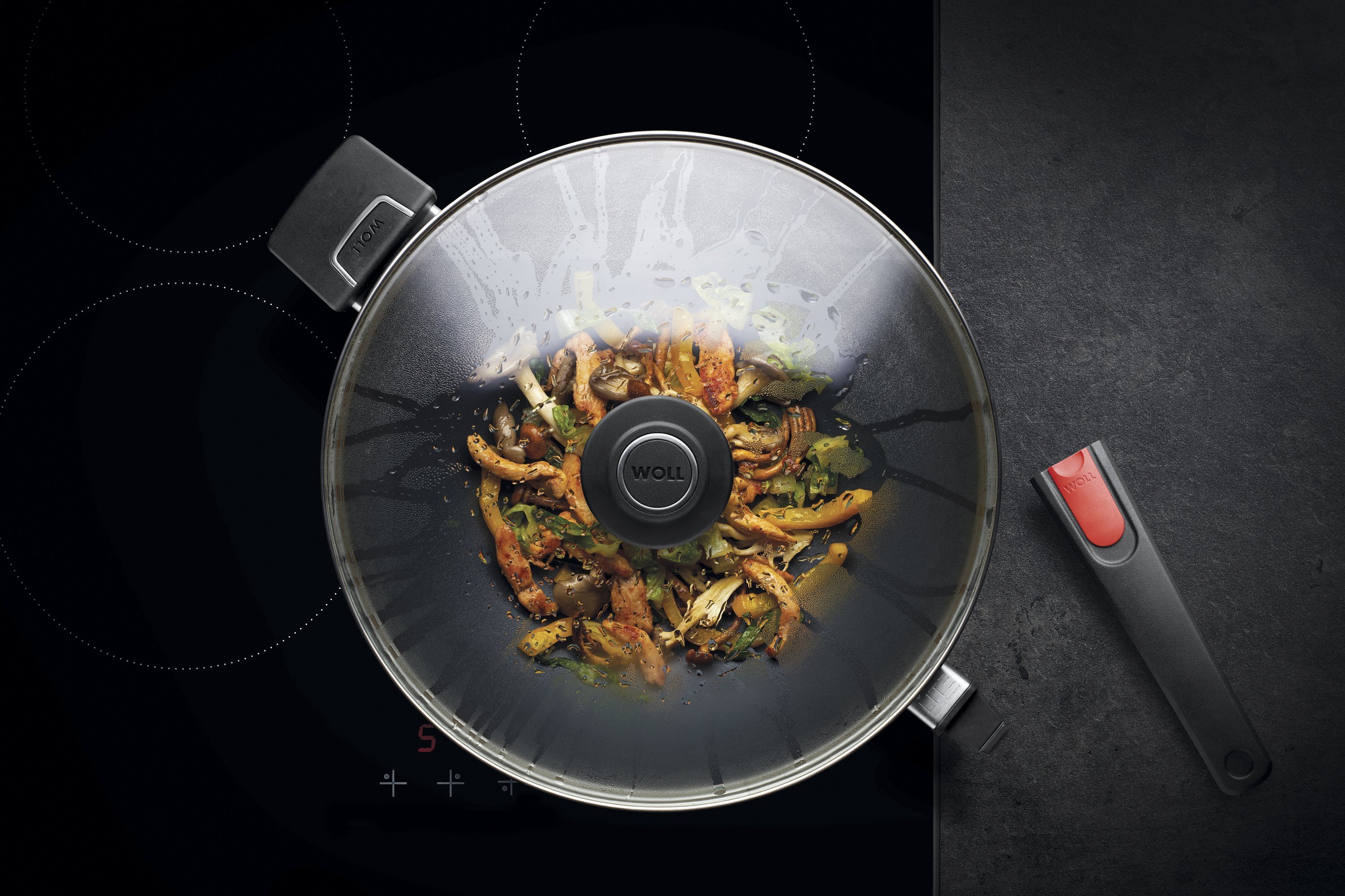 Auf dem Herd steht eine runde Diamond Lite Wok- und Rührpfanne  In dieser befindet sich ein  frisch zubereitetes Gericht. Am Glasdeckel hat sich Kondenswasser gebildet.