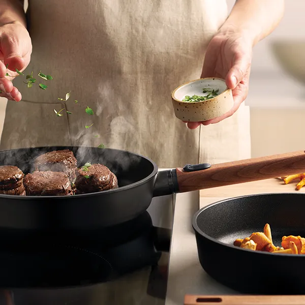 Auf dem Herd steht eine runde, heiße Pfanne der Serie Eco Logic QXR mit abnehmbarem Holzstiel, in der Fleisch angebraten und mit Kräutern verfeinert wird. Rechts daneben steht eine weitere Pfanne derselben Serie mit abgenommenem Holzstiel. In der Pfanne s