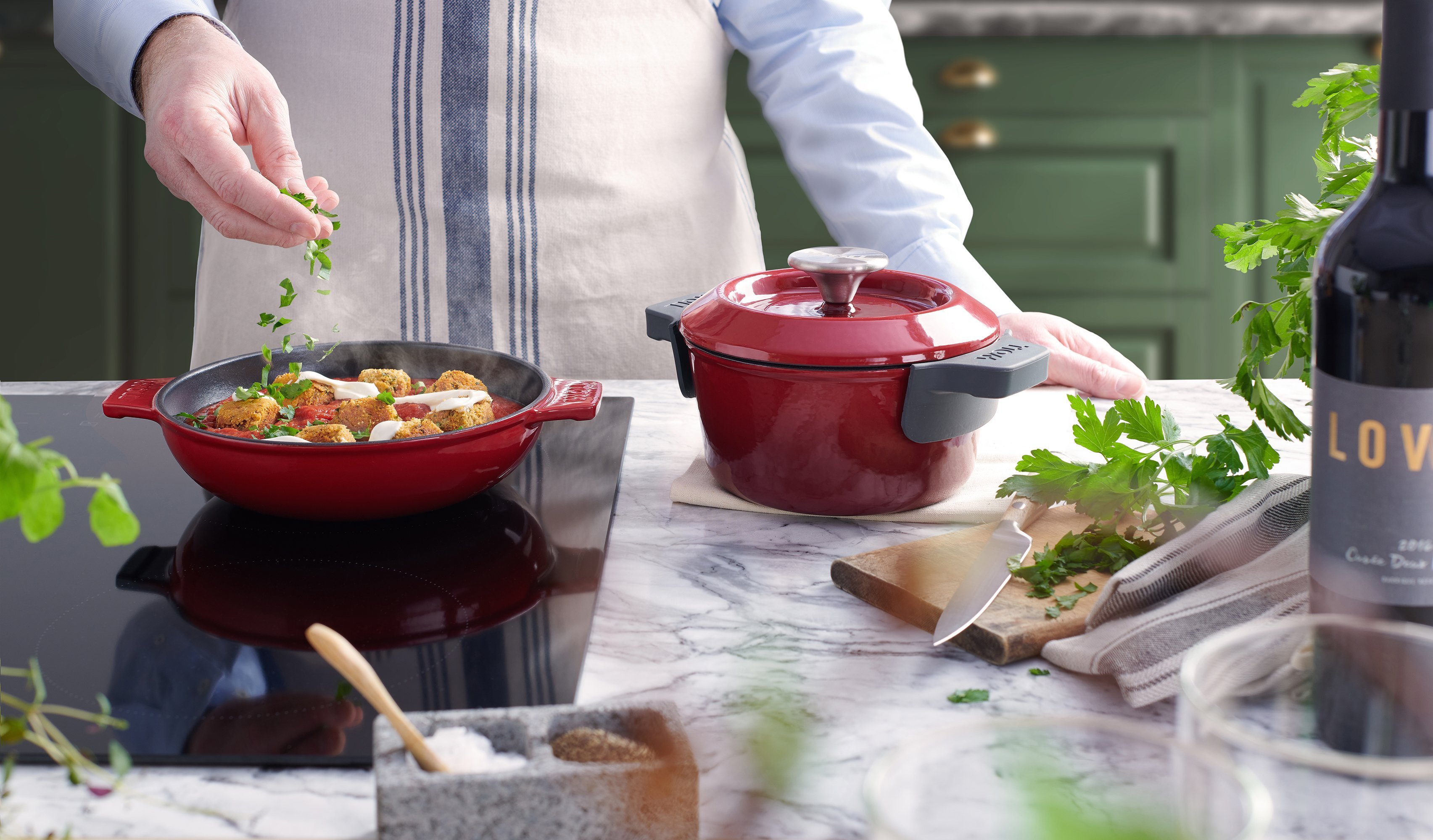 Auf dem Herd steht eine rote, runde Iron Servierpfanne, in der sich ein dampfendes Gericht befindet. Daneben steht ein Iron Topf mit Deckel.