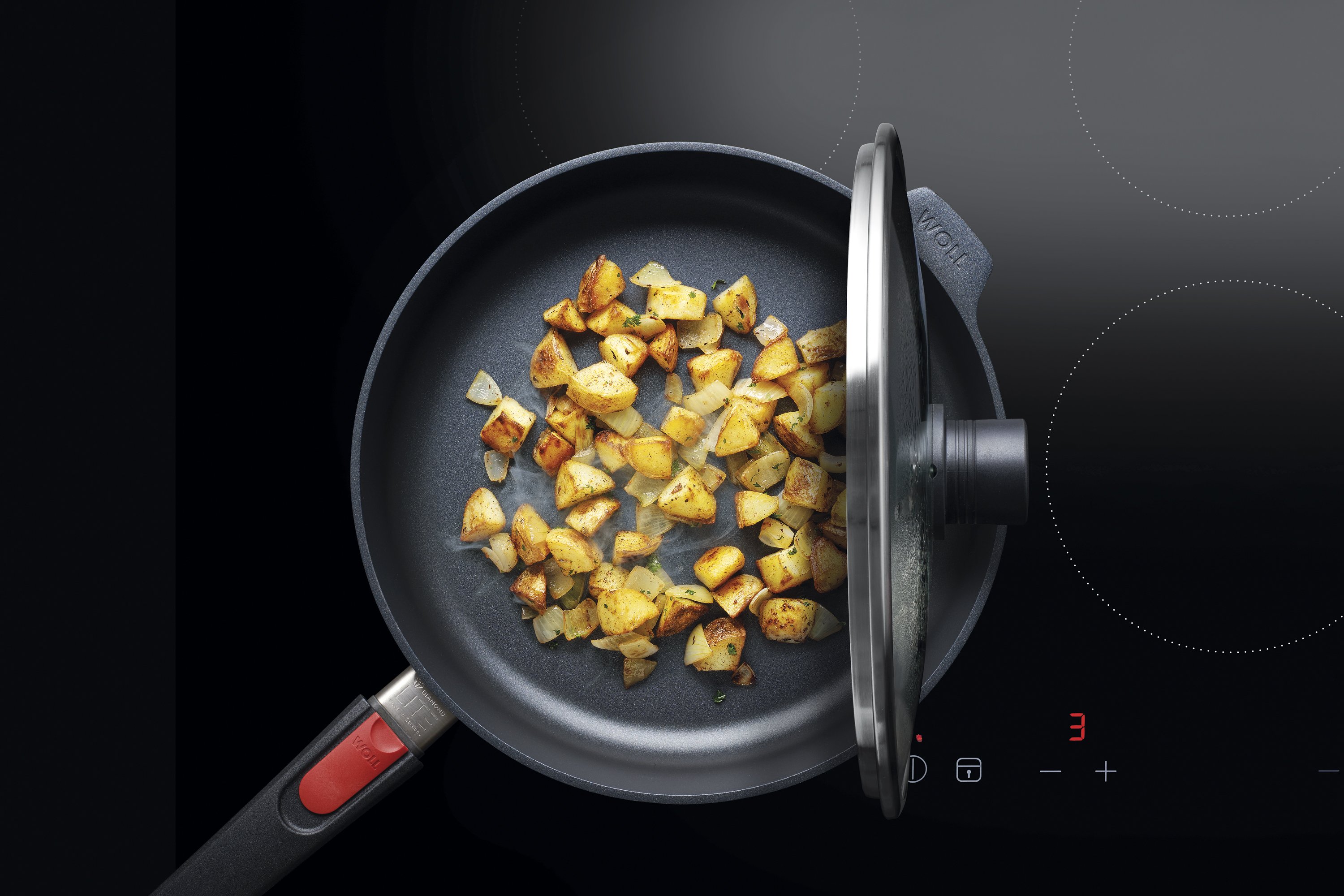 Auf dem Herd steht eine runde, heiße Pfanne aus der Serie Diamond Lite mit abnehmbarem Stiel. In dieser befindet sich ein  frisch zubereitetes Gericht. Der Glasdeckel ist mit der verlängerten Griffseite der Smart-Lid-Serie an der Kante der Pfanne eingehän