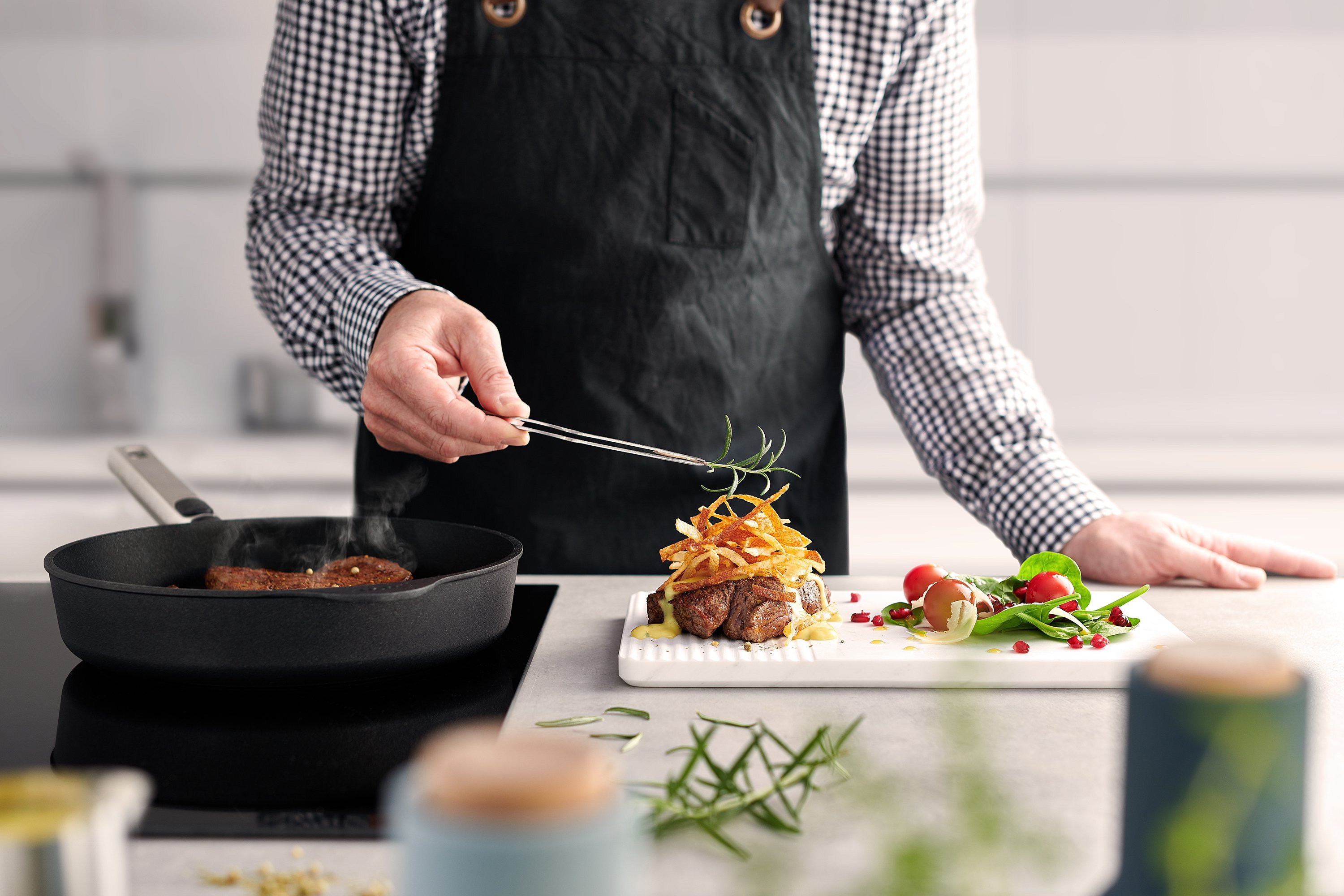 In einer runden Logic XR Pfanne wird Fleisch angebraten. Daneben wird Fleisch auf einem Brett garniert. 