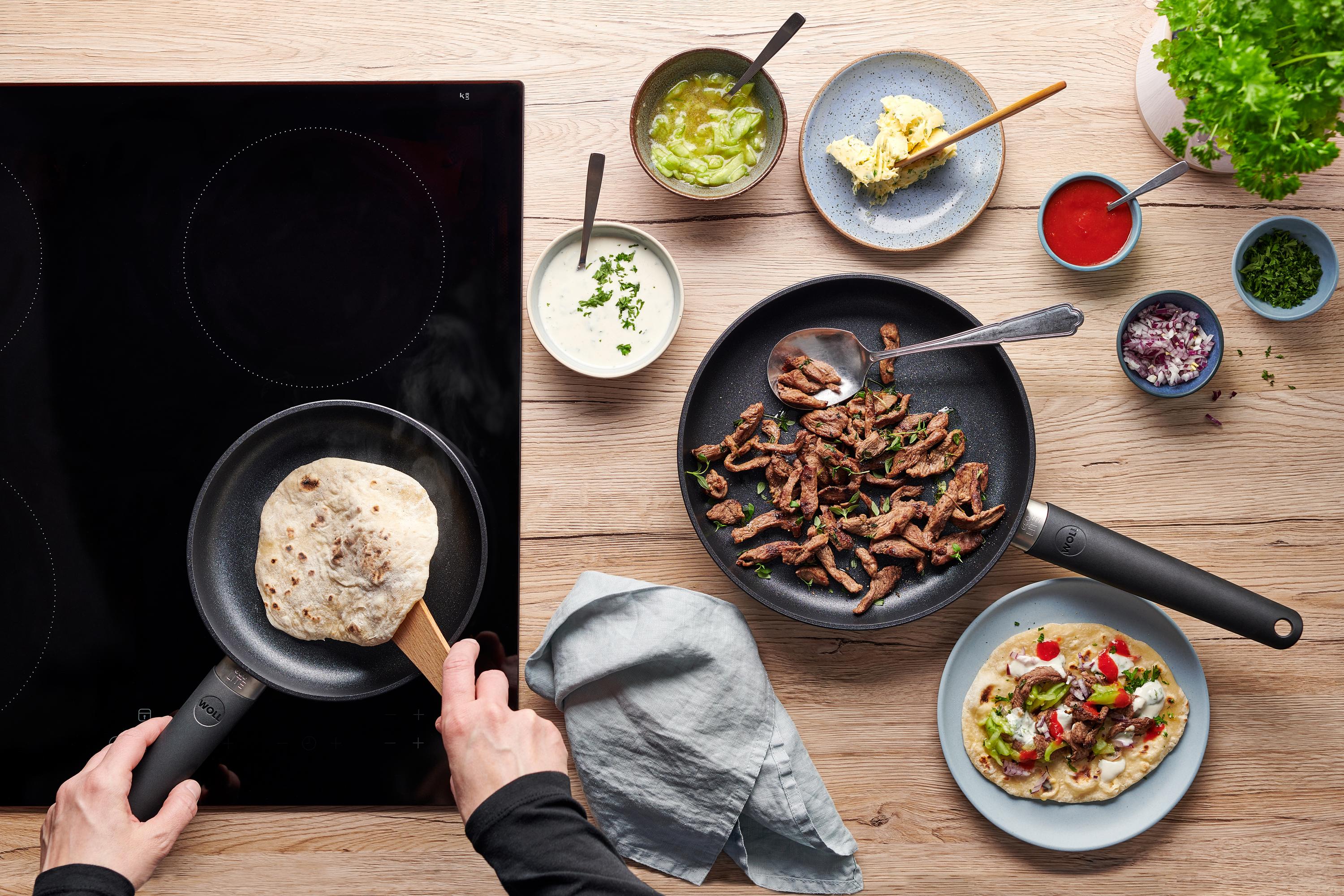 In der runden Eco-Lite-QXR-Pfanne werden Fladenbrote gebacken. Daneben liegt in einer Pfanne angebratenes Lamm und auf einem Teller ist Döner zubereitet.