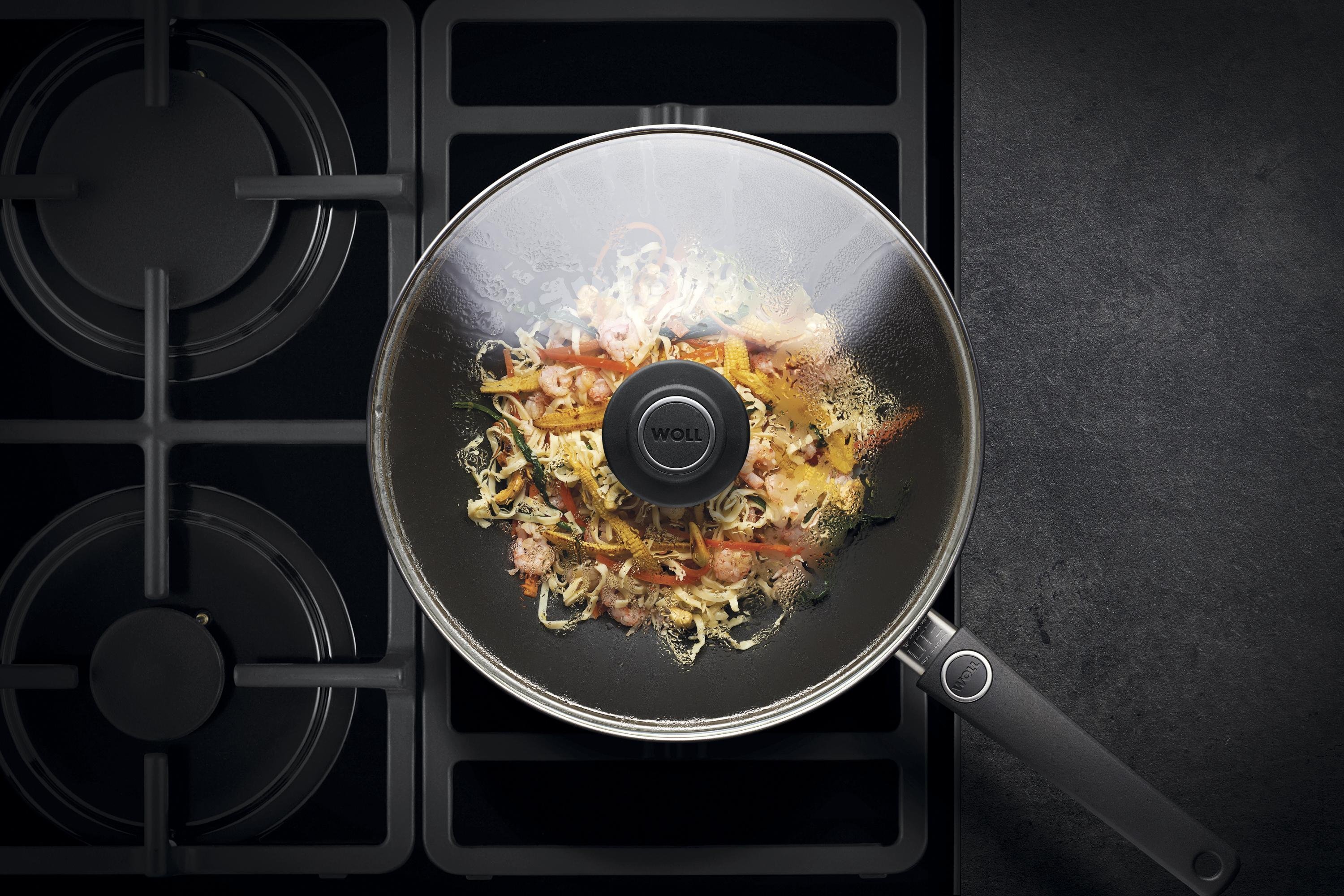 Auf dem Herd steht eine runde Diamond Lite Wok- und Rührpfanne  In dieser befindet sich ein  frisch zubereitetes Gericht. Am Glasdeckel hat sich Kondenswasser gebildet.