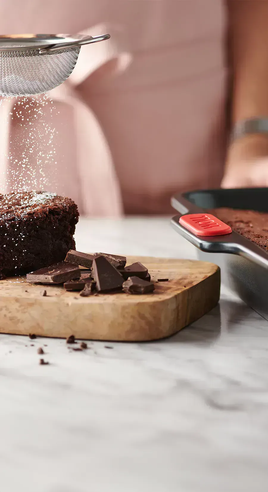 Auf einem Holzbrett liegt ein frisch gebackenes Stück Schokoladenkuchen, das mit Puderzucker bestäubt ist. Schokoladenstückchen auf dem Holzbrett dienen als Dekoration. Daneben steht die rechteckige Backform der Let's Bake!-Serie, die mit weiterem Schokol