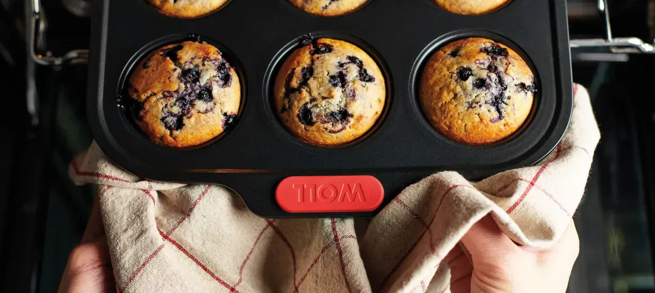 Eine heiße rechteckige Muffinform der Serie Let's Bake! enthält frisch gebackene Blaubeermuffins. Sie wird mit einem Küchentuch aus dem Backofen genommen.