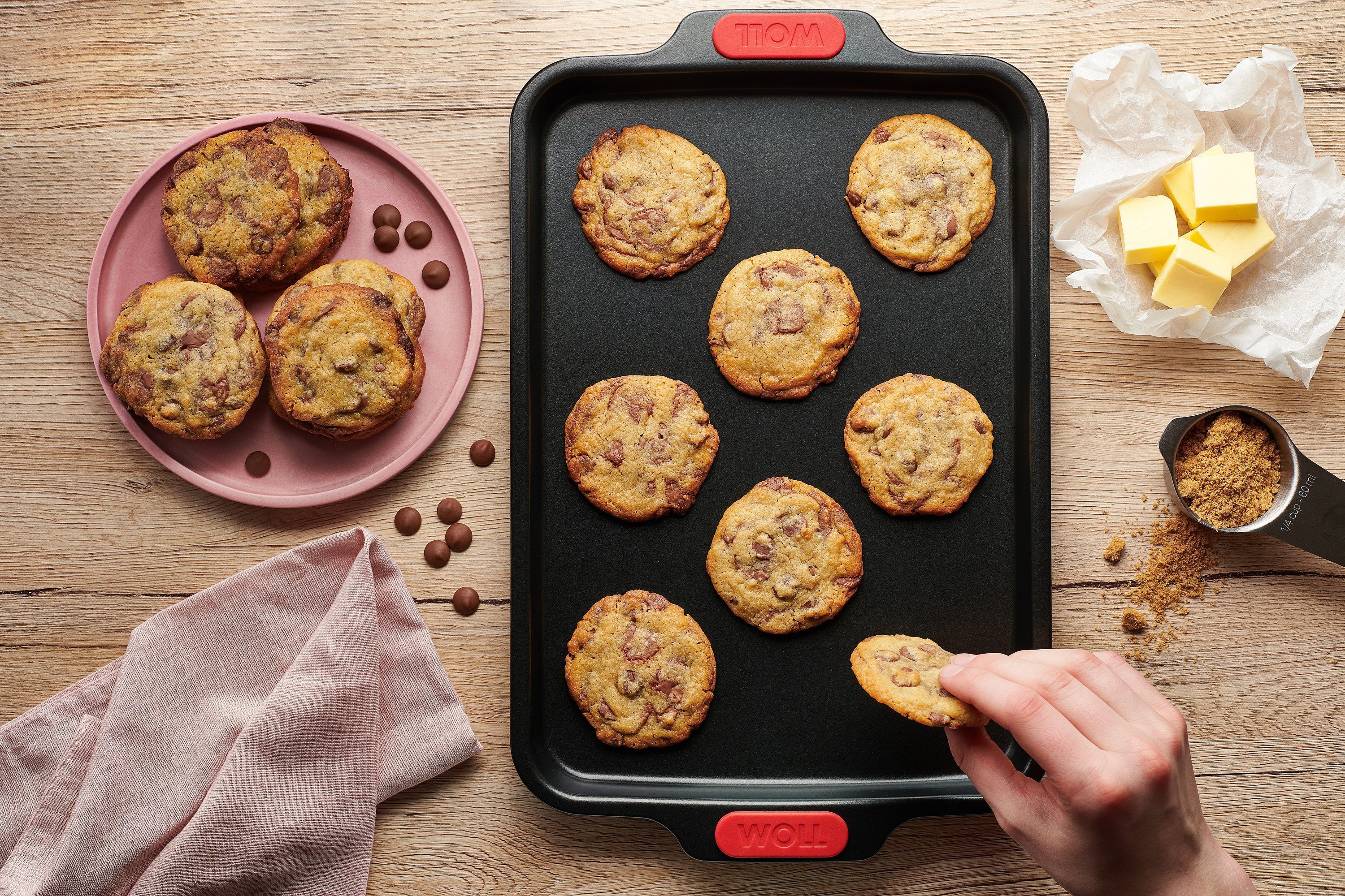 Auf einem rechteckigen Let's bake! Backblech liegen frisch gebackene Cookies mit Schokoladestückchen. Ein Cookie wird vom Blech genommen.