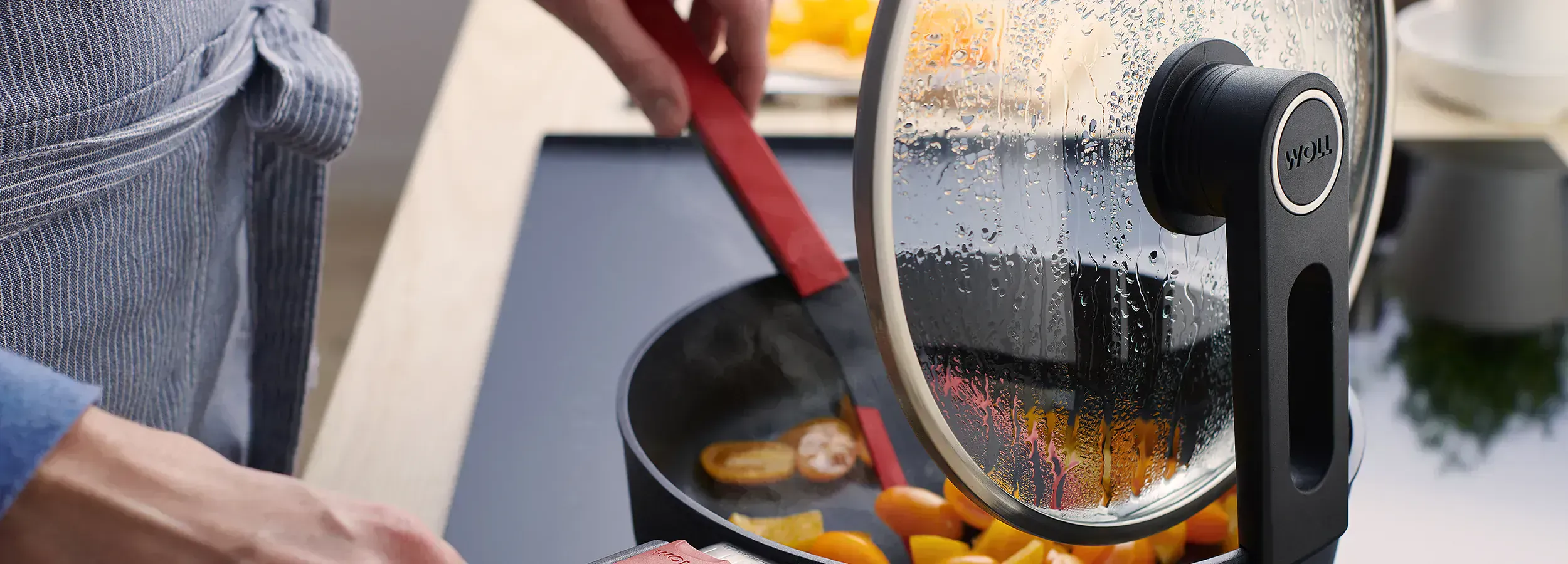 In einer heißen Diamond-Lite-Pfanne wird gerade ein dampfendes Gericht mit einem Cock-it-Pfannenwender umgerührt. Der Glasdeckel ist mit der verlängerten Seite der Smart-Lid-Serie an der Kante der Pfanne eingehängt.