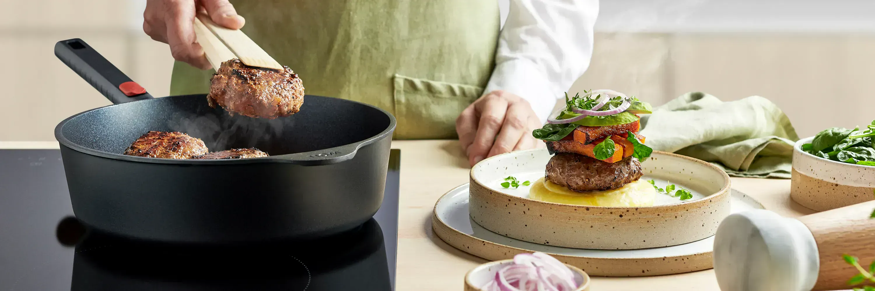In einer runden Hochrandpfanne mit festem Stiel der Serie Eco Logic QXR liegt fertig angebrachtes Fleisch. Daneben steht ein Teller mit Kartoffelpüree, darauf ein Stück Fleisch, garniert mit angebraten Karotten und etwas Salat.