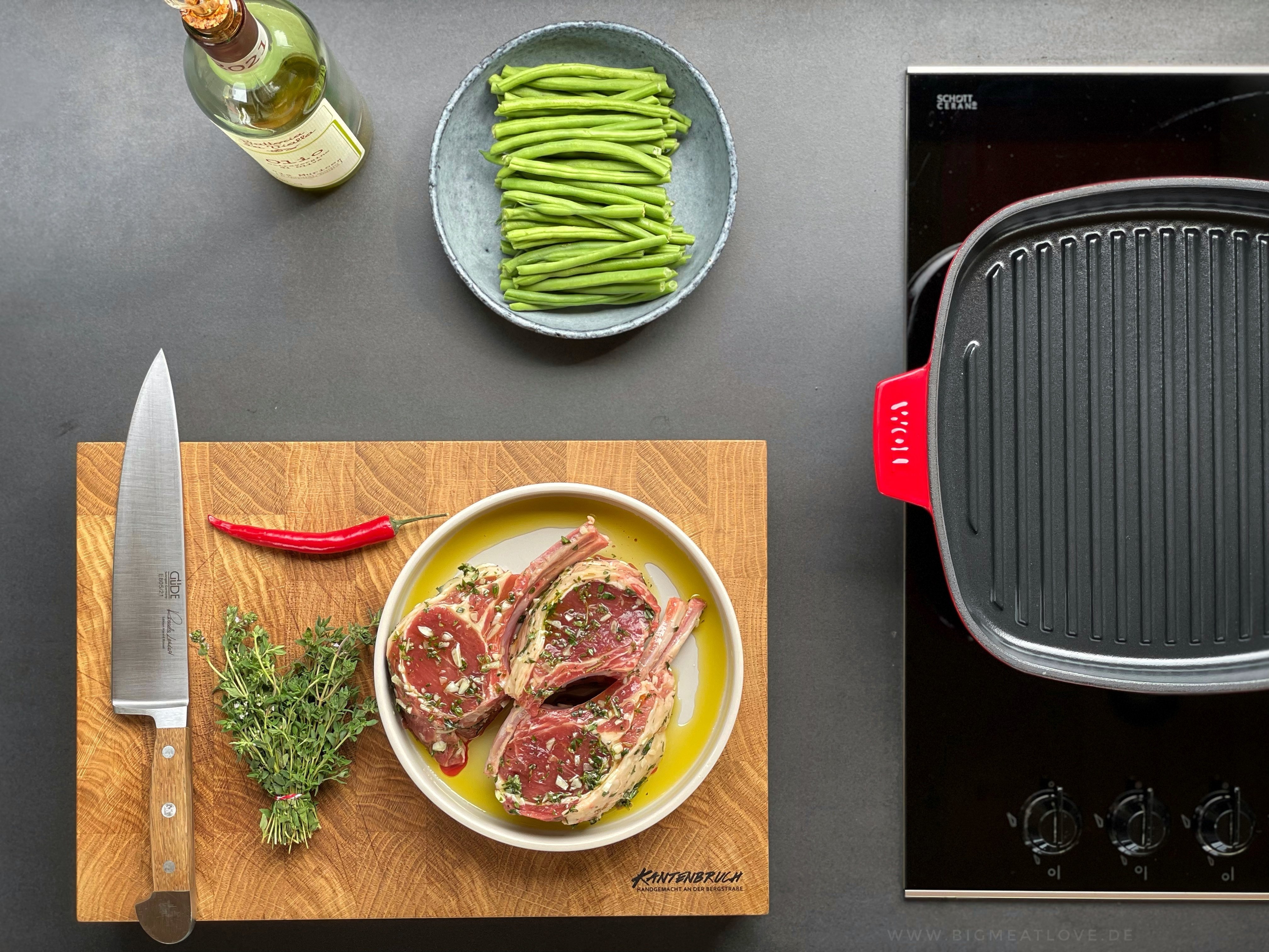 Auf dem Herd steht eine viereckige, rote Iron Grillpfanne mit Rillen.  Auf einem Teller daneben liegt mariniertes Fleisch. 