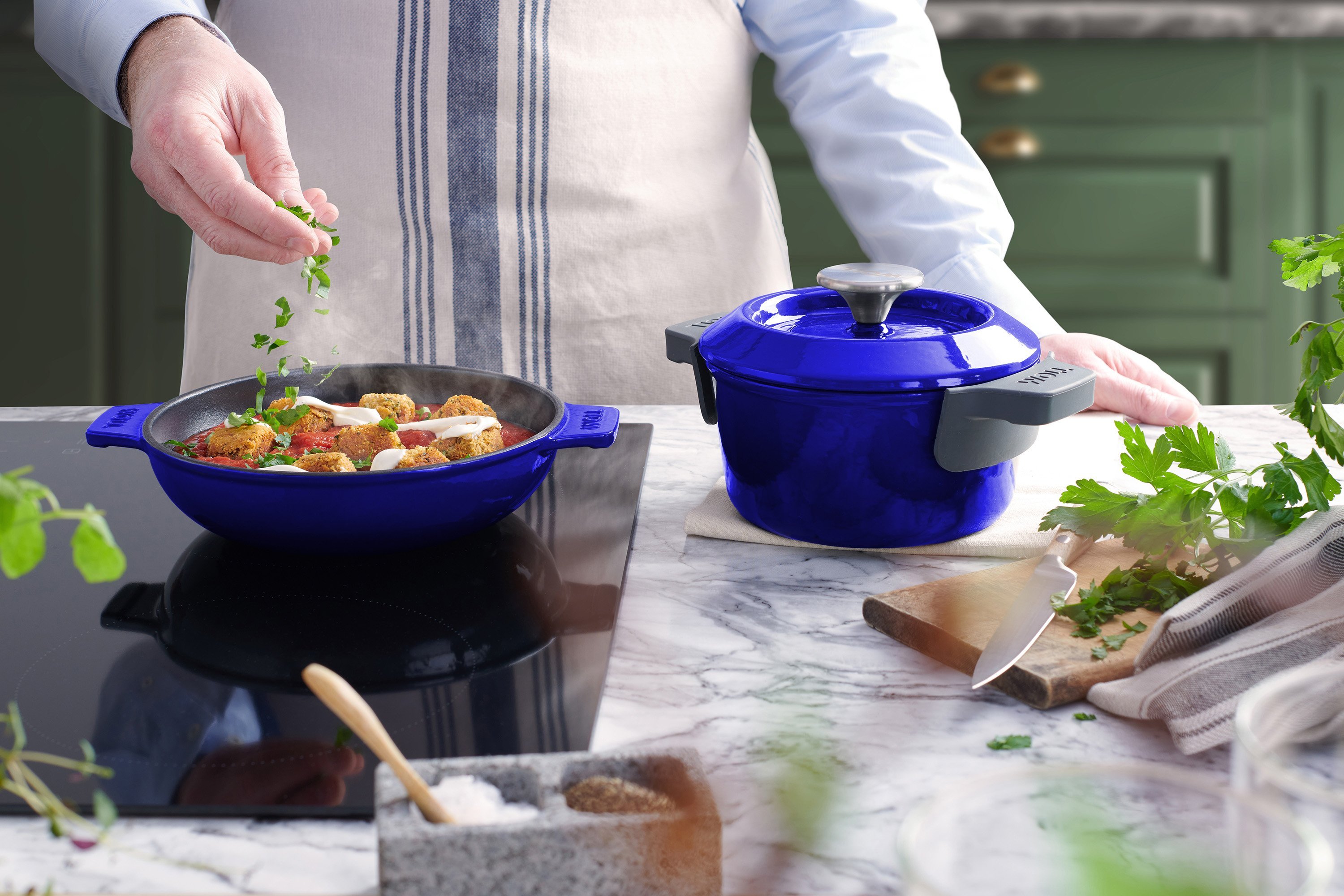 Auf dem Herd steht eine blaue, runde Iron Servierpfanne, in der sich ein dampfendes Gericht befindet. Daneben steht ein Iron Topf mit Deckel.