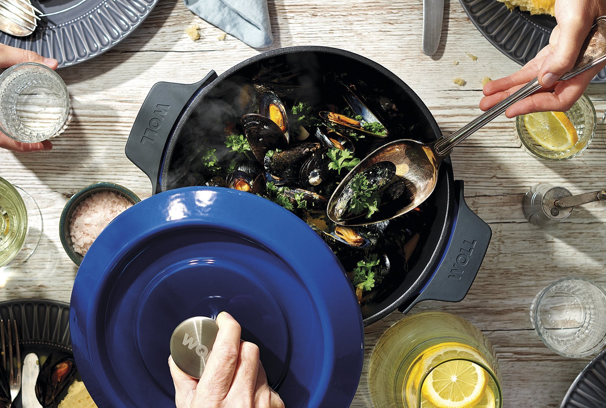 Auf dem Tisch steht eine runde, blaue Iron-Kasserolle mit zubereiteten Muscheln. Der Deckel wird hochgehoben.