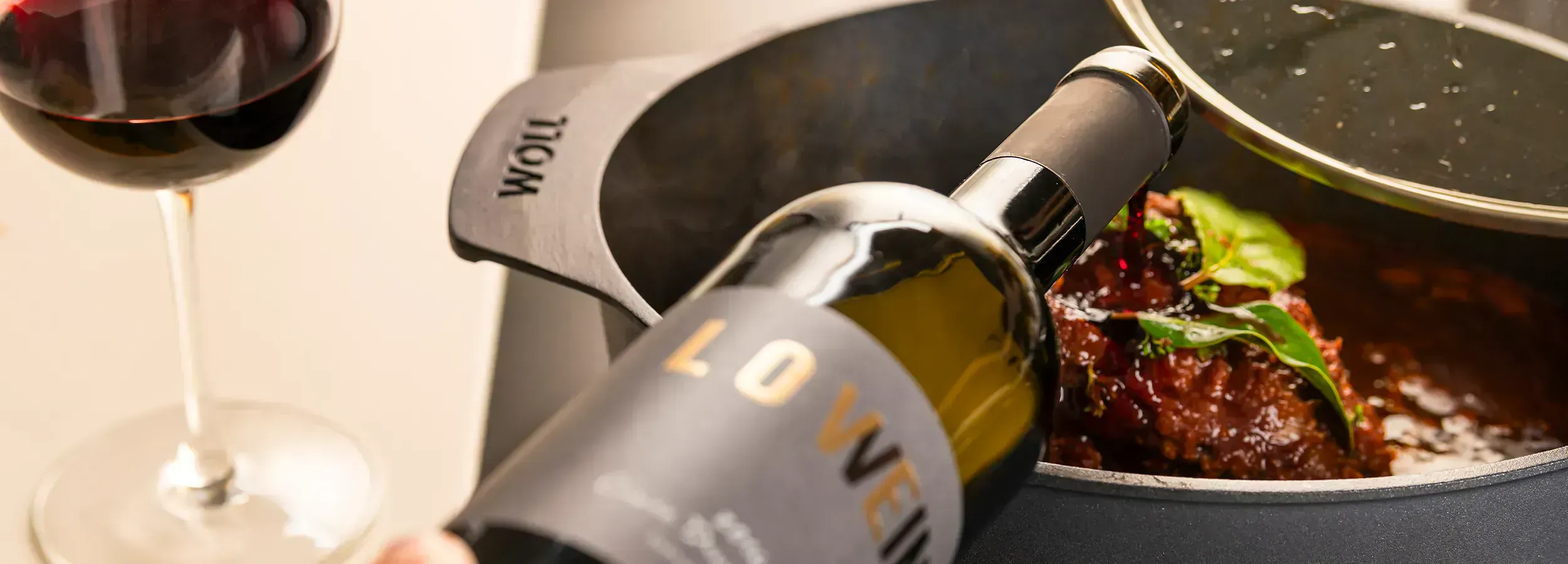 Ein heißer Bräter der Serie Diamond Lite enthält ein Gericht. Dieses wird mit Rotwein übergossen. Ein Rotweinglas ist daneben zu sehen.