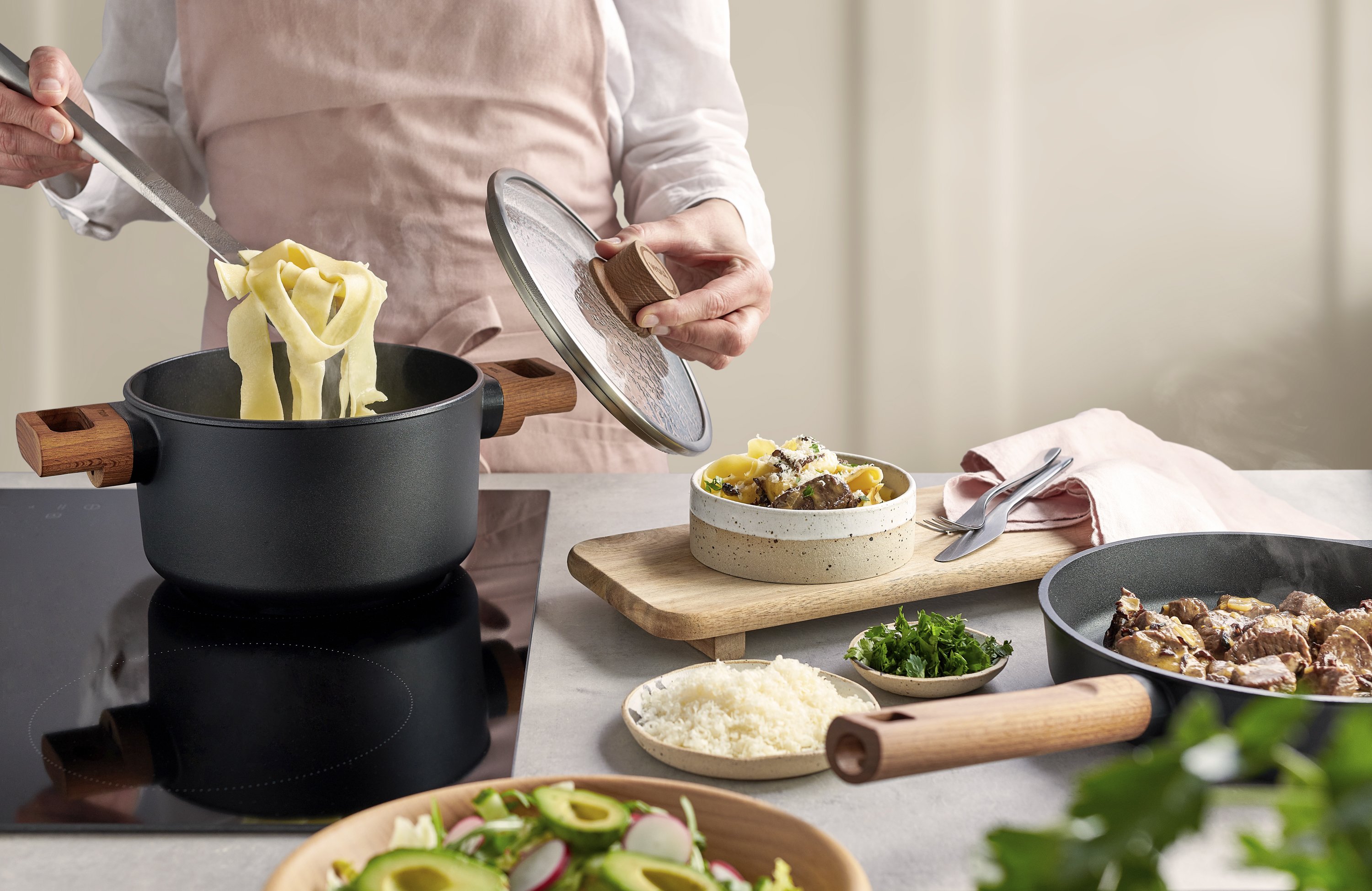 Im Topf der Serie Eco Logic QXR wurden frische Nudeln gekocht, die nun entnommen werden. Dazu wurde der Deckel abgenommen. Auf der Arbeitsplatte steht ein Holzbrett mit einem angerichteten Pastateller. Daneben liegen Besteck und eine Serviette. Um den Tel