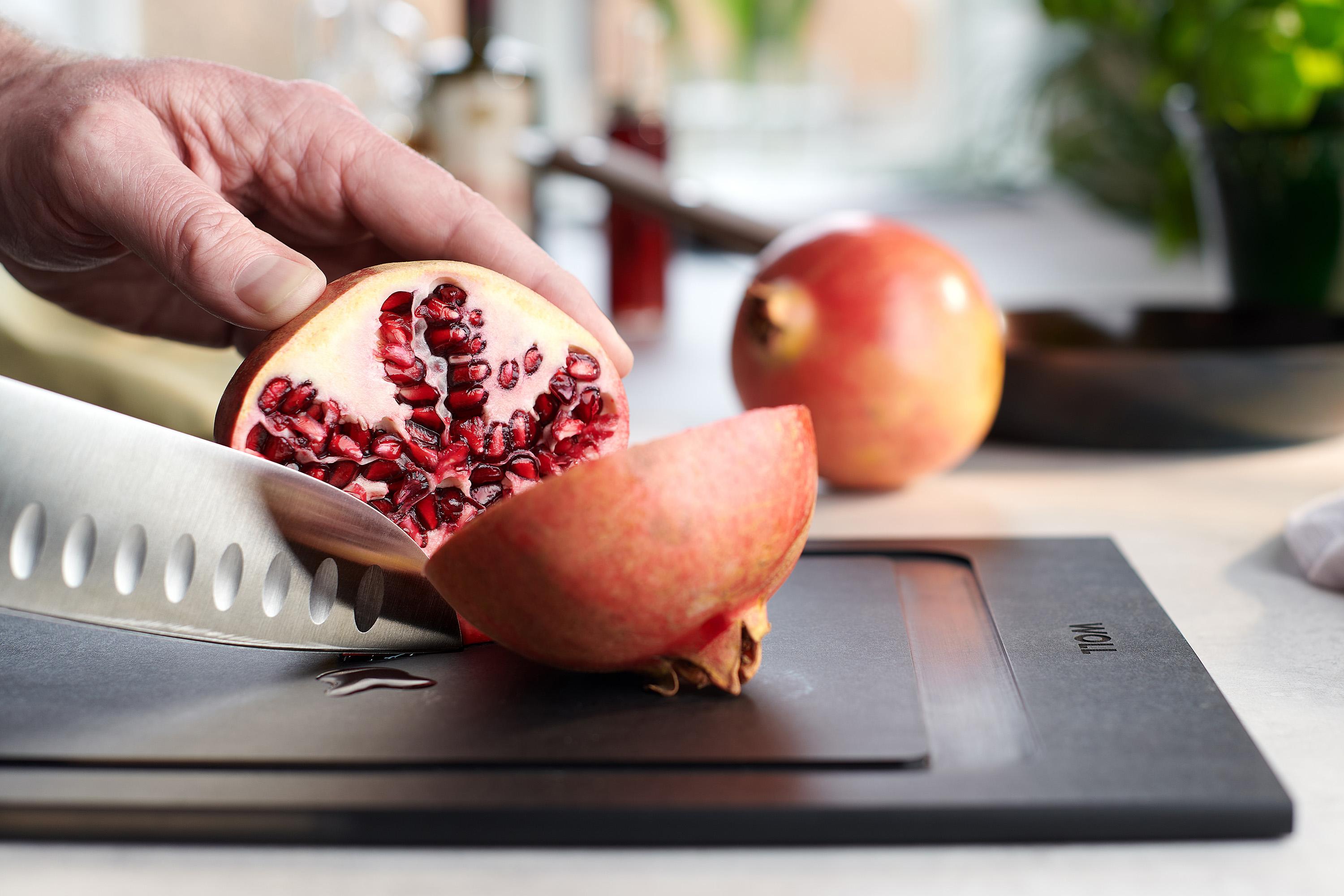 Ein Granatapfel wird auf einem Cut & Serve Schneidebrett  mit einem Messer geteilt.